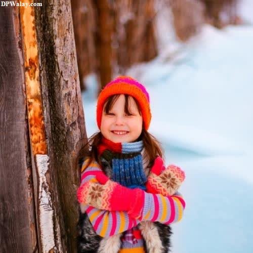 a little girl in a colorful sweater and hat standing by a tree