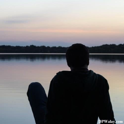 a man sitting on the edge of a lake looking out at the sunset 