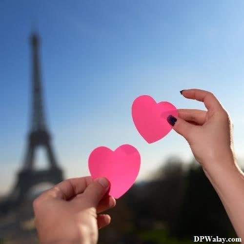 two hands holding pink paper hearts in front of the eiff tower