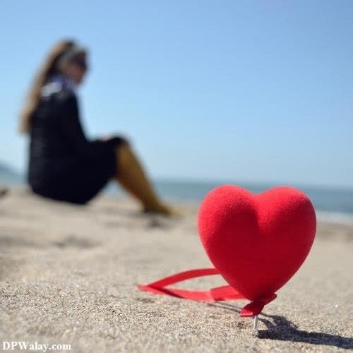 a red heart on the beach with a woman sitting in the background love dp pic hd